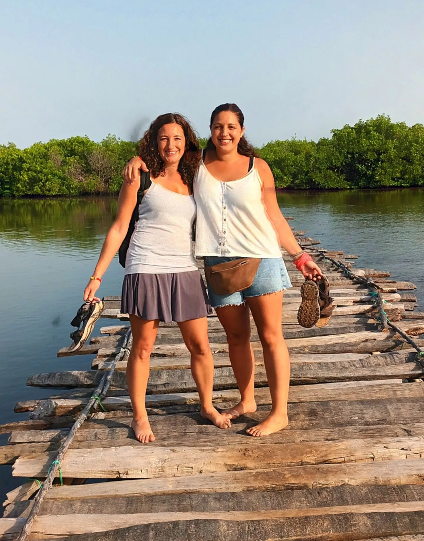 Meri y Núria, de Alas en la Mochila, sonríen mientras caminan descalzas sobre un puente rústico de madera en un manglar de Senegal.